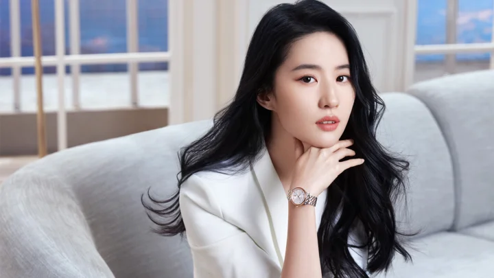 검은 머리의 여성이 흰색 상의를 입고 밝은 창문을 배경으로 연한 색 소파에 앉아 손을 턱 가까이에 대고 있다.