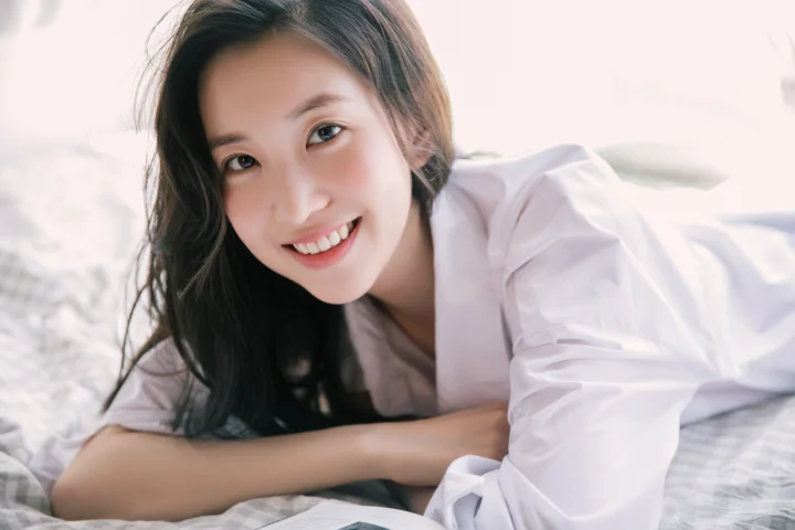 白いシャツを着た成人女性が、ベッドに横たわりながらカメラに笑顔を向けています。
