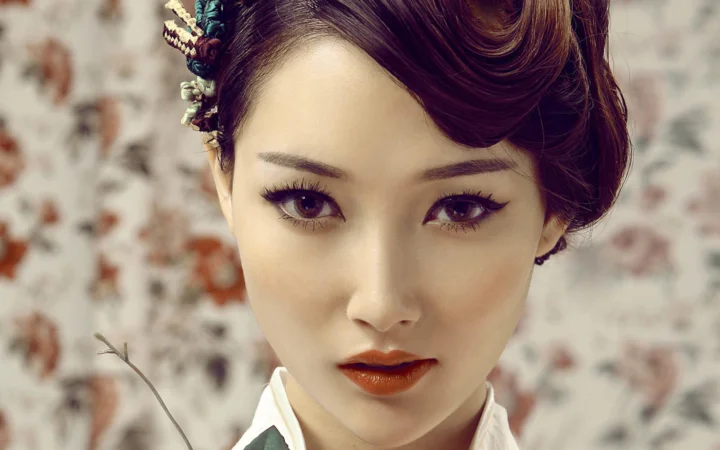 一位成年女性的近距离肖像，留着盘发并化着复古妆容，背景为柔和的花纹墙面。