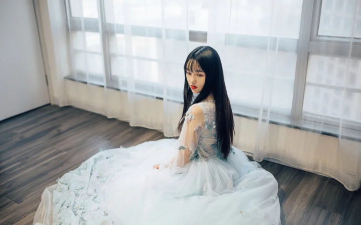 흰색 드레스를 입은 여성이 밝은 창가의 실내 바닥에 앉아 있습니다.