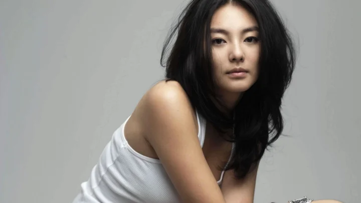 一名黑发女性穿着白色背心，在灰色背景前面向镜头。