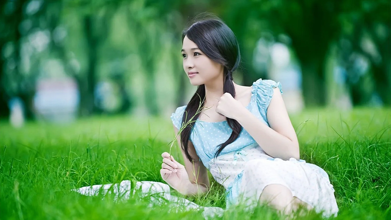 女性が緑の草地に座り、背後に木々と柔らかくぼけた屋外の背景が広がっています。