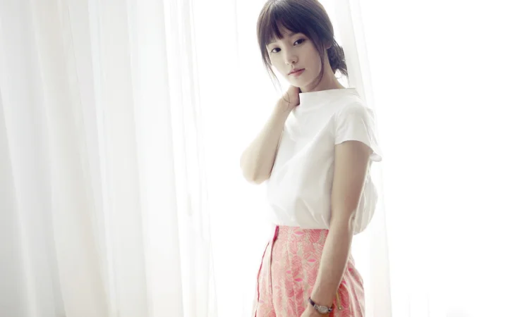 흰색 상의를 입은 여성이 밝은 커튼 옆에 서 있다.