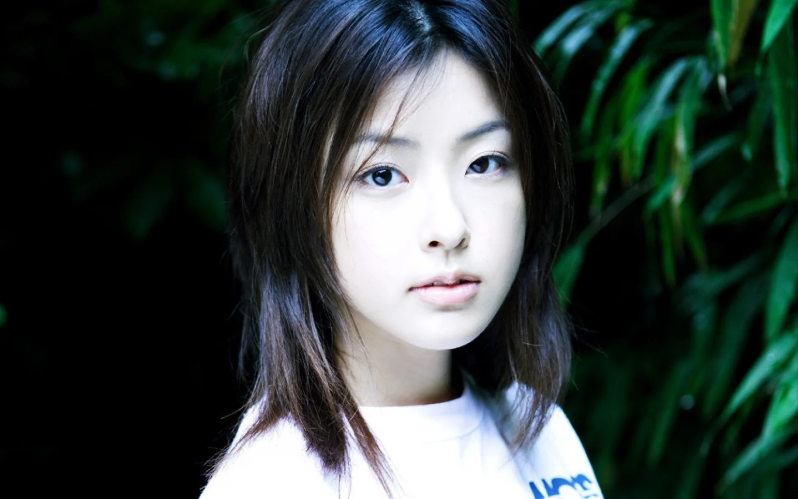 검은 머리의 여성이 흰색 상의를 입고 짙은 녹색 나뭇잎을 배경으로 카메라를 바라보고 있습니다.