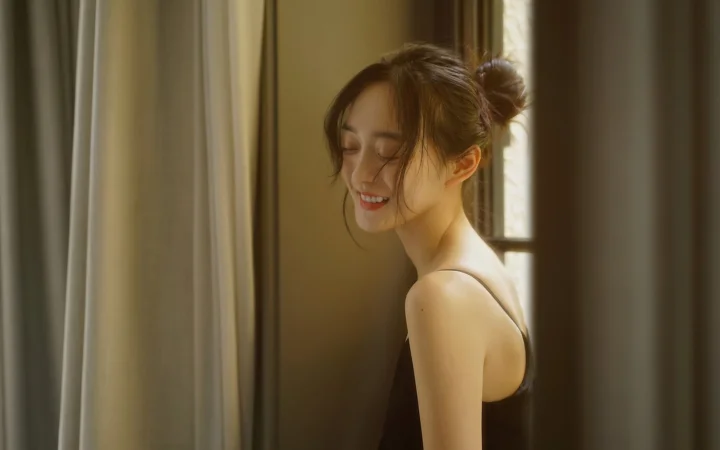一名女性站在窗边和窗帘之间，沐浴在温暖的光线中。