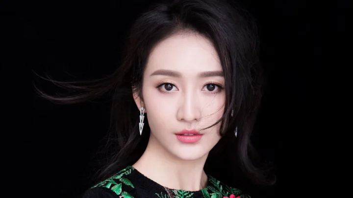 黑色背景前，一位深色长发女性直视镜头，佩戴银色耳饰并穿着带绿色图案的黑色上衣。