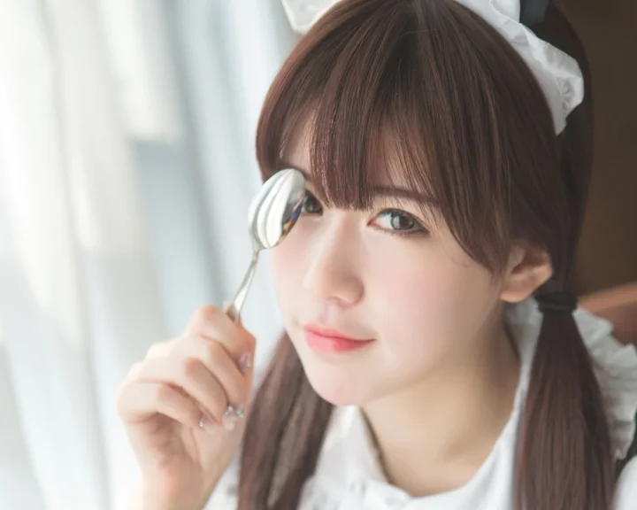 갈색 머리 여성이 흰색 헤드드레스를 착용하고 부드러운 실내 조명 속에서 얼굴 가까이에 숟가락을 들고 있다.