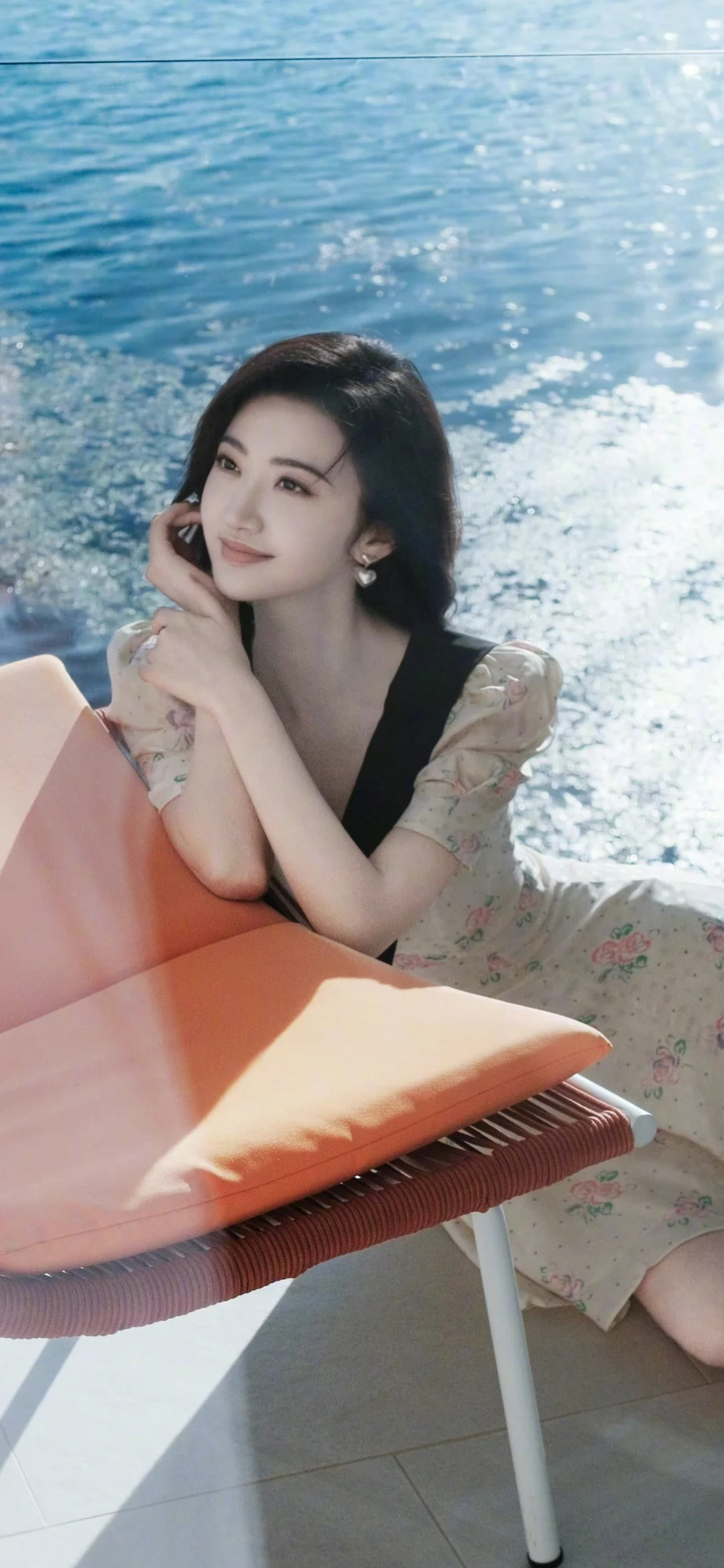 一名成年女子坐在橙色椅子上，身后是阳光下闪耀的水面。