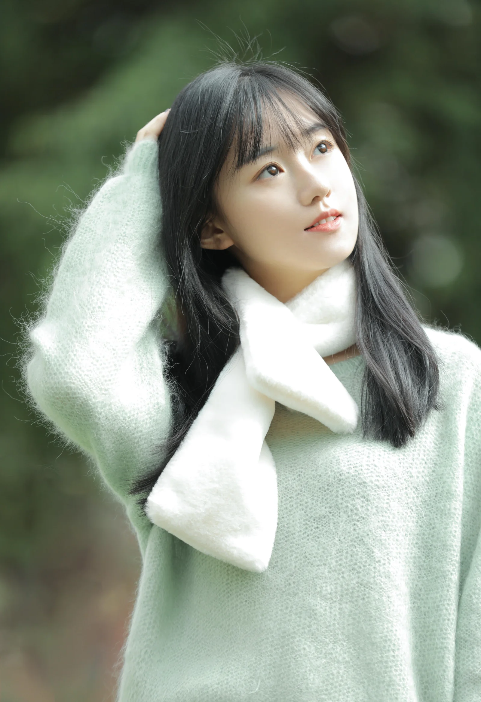 長い髪の女性がミントグリーンのセーターと白いマフラーを身に着け、緑にぼけた屋外の背景で上を見つめている。