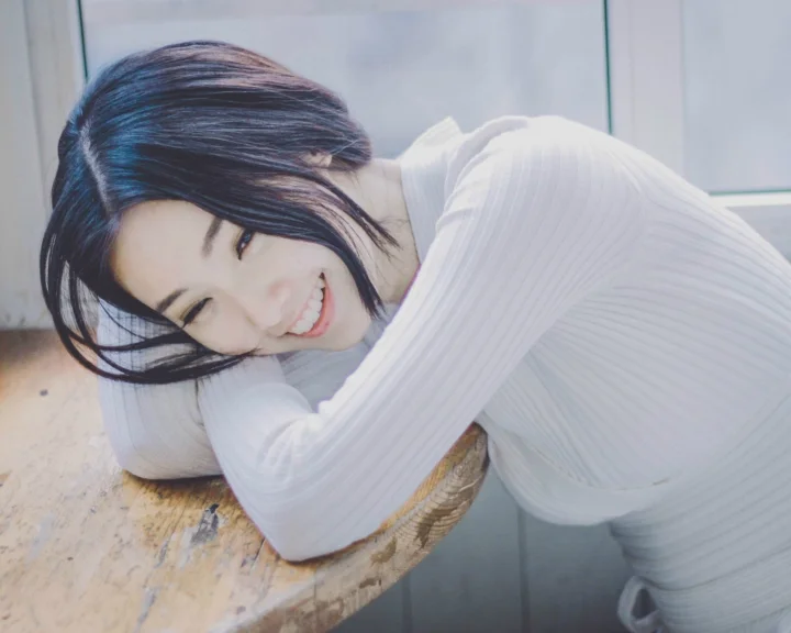 一名穿白色长袖上衣的女子俯身靠在木桌旁微笑。