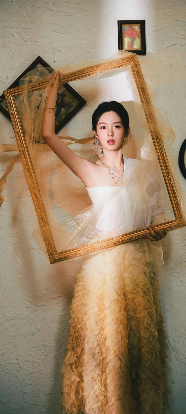 一名身穿白色礼服的成年女性手持金色画框站在墙前。