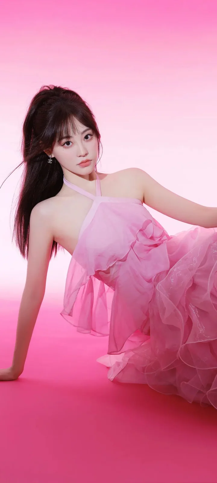 一位成年女性身穿粉色连衣裙，在粉色背景前侧身坐姿面向镜头。