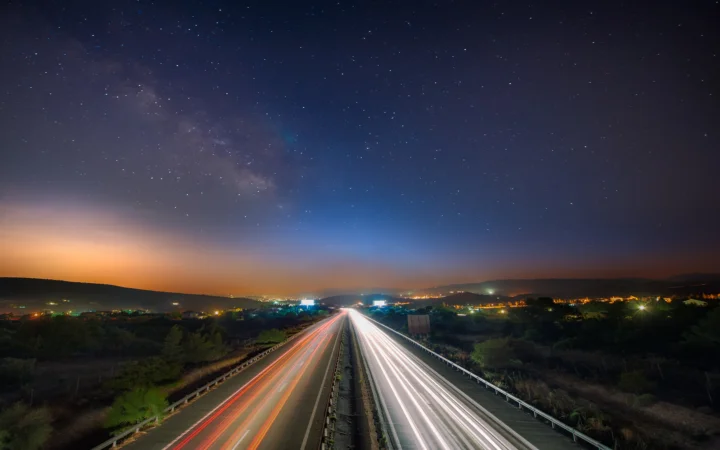 夜空中的银河横跨公路上方，远处城镇灯光与车流光轨延伸至地平线。
