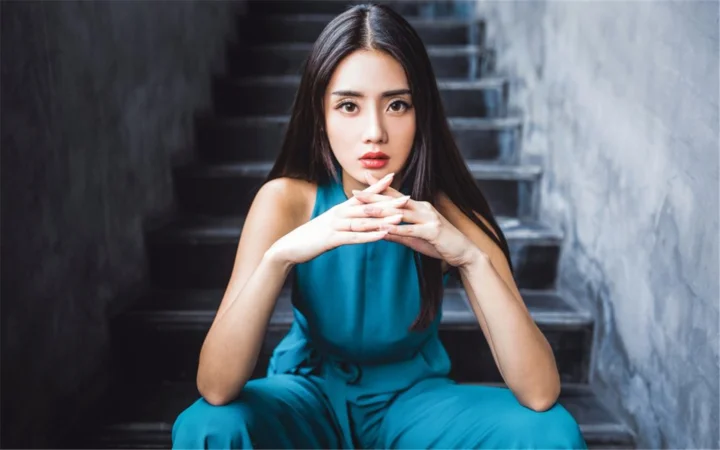 一名身穿蓝色无袖服装的女子坐在楼梯上，双手交叠置于下巴前方。
