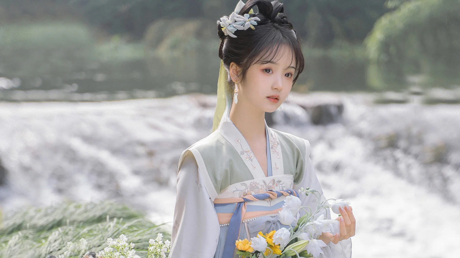 전통 의상을 입은 여성이 꽃을 들고 폭포 앞에 서 있다.