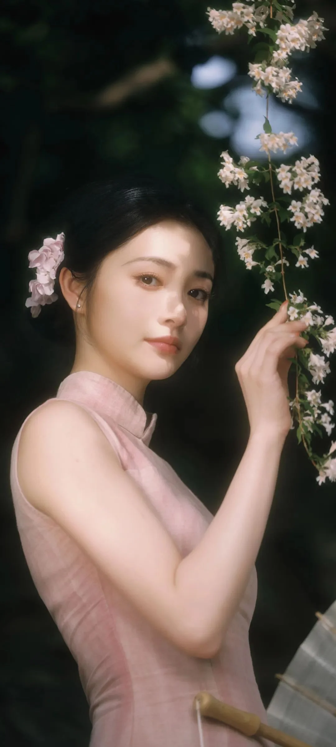연한 분홍색 민소매 옷을 입은 여성이 어두운 배경 앞에서 옆의 꽃이 핀 가지를 만지고 있다.