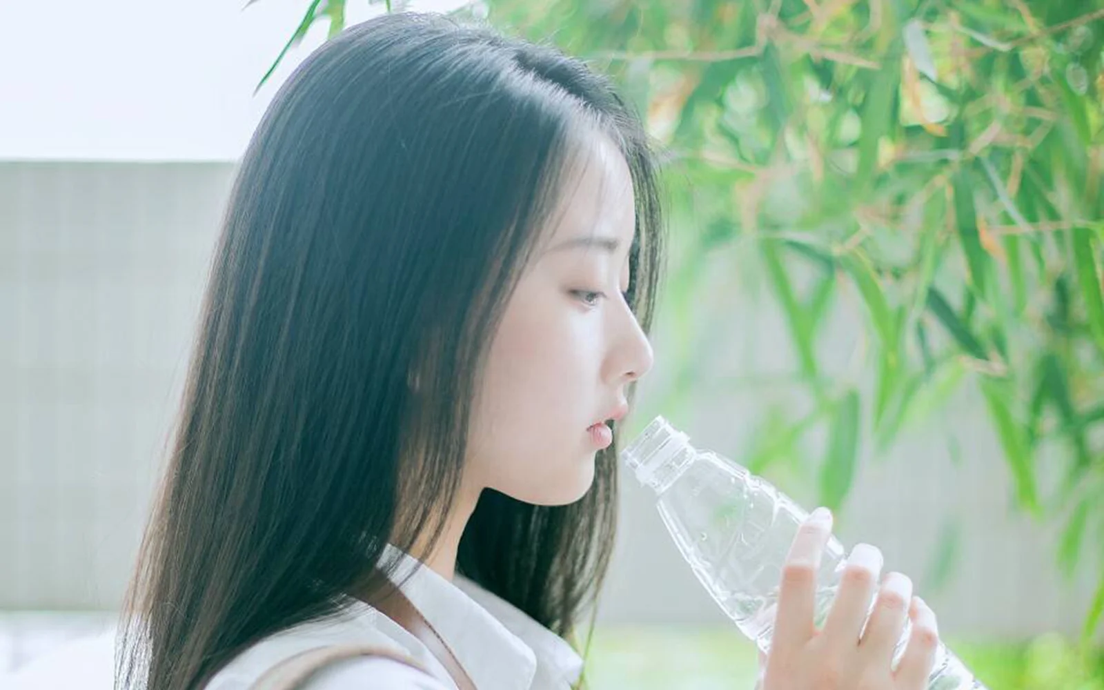 창가에서 여성이 투명한 물병으로 물을 마시고 옆에 초록색 대나무 잎이 보입니다.