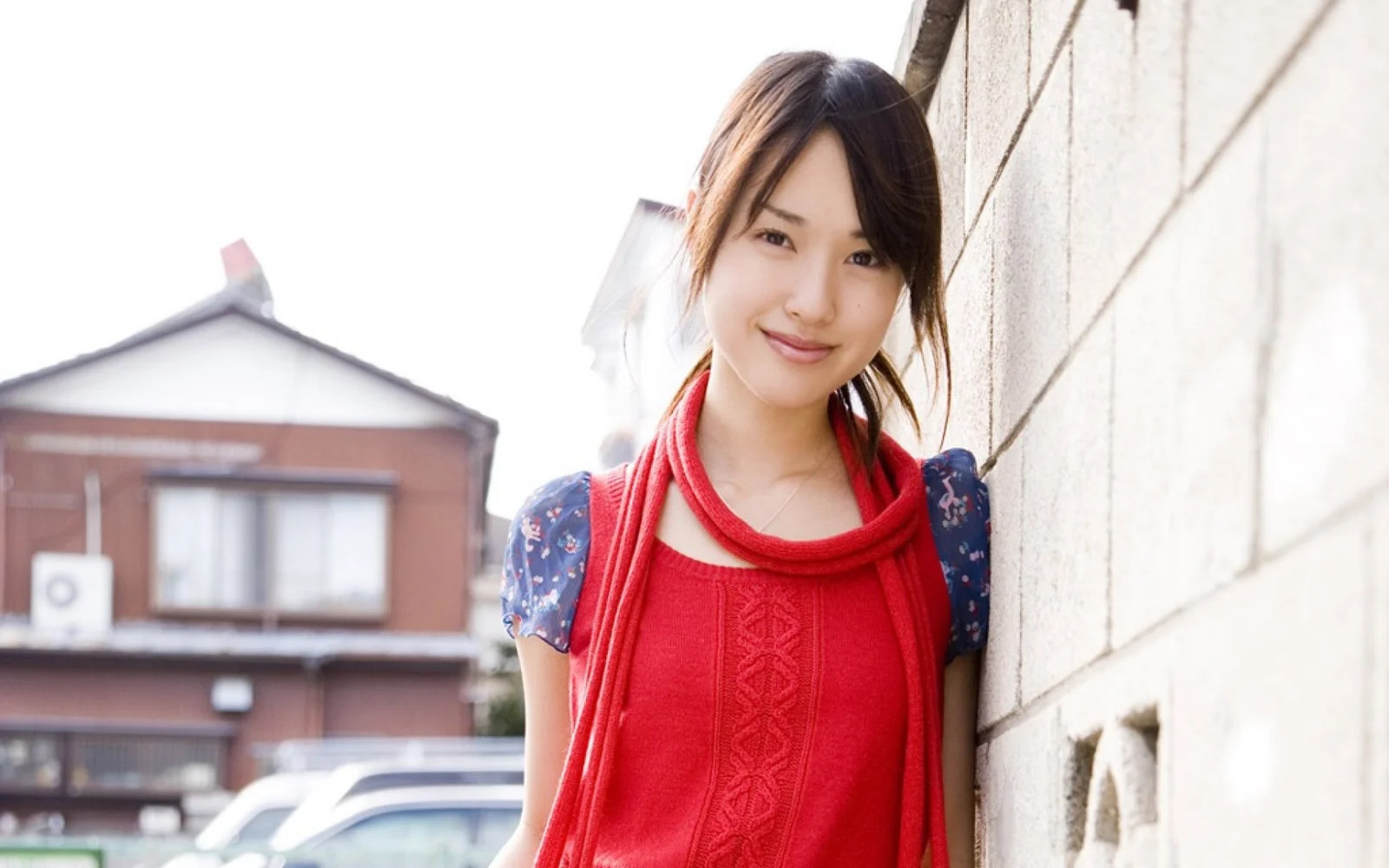 一名佩戴红色围巾的女子站在户外砖墙旁微笑面对镜头。