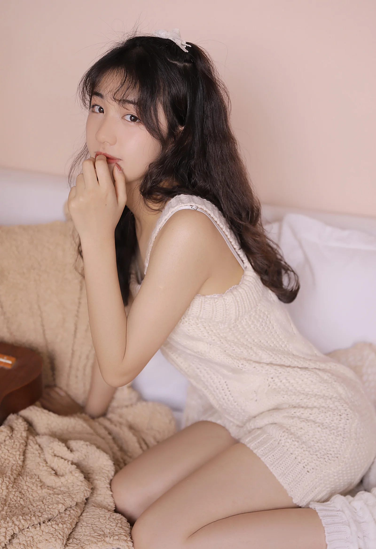 淡い色のニットワンピースを着た女性がベッドに座り、カメラに向かってポーズを取っている。