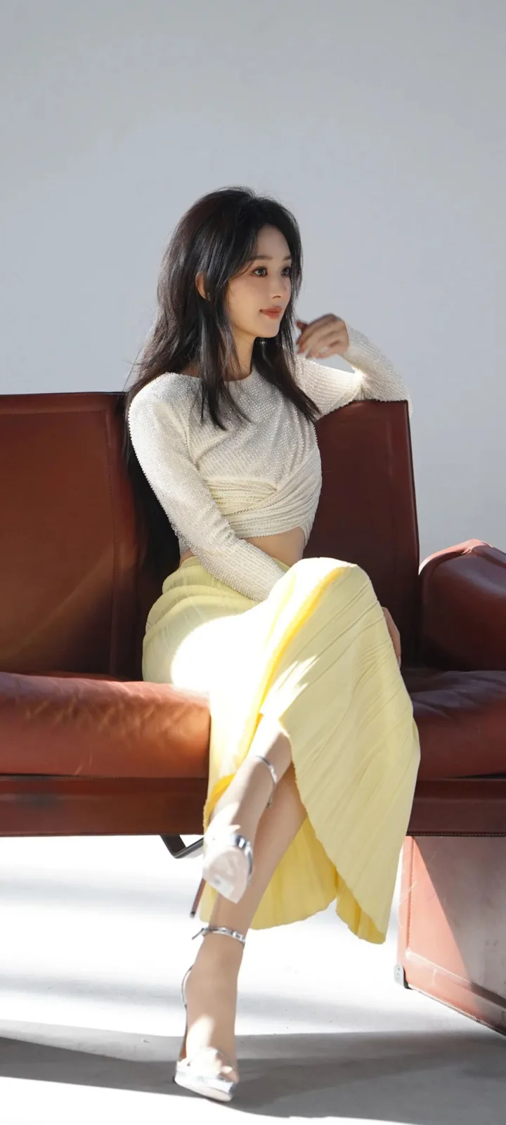 一名女子穿着白色上衣和浅黄色裙子，坐在棕色皮沙发上，阳光照亮了室内空间。