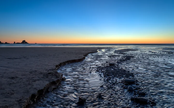 暮色中的海岸线上，浅滩水流穿过深色卵石与湿润沙地，地平线被橙色余晖映亮。