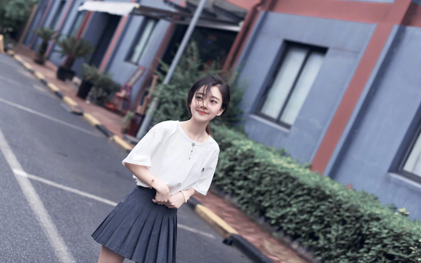 一名身穿白色上衣和深色裙子的女性站在城市街道上，身后是建筑和绿植。