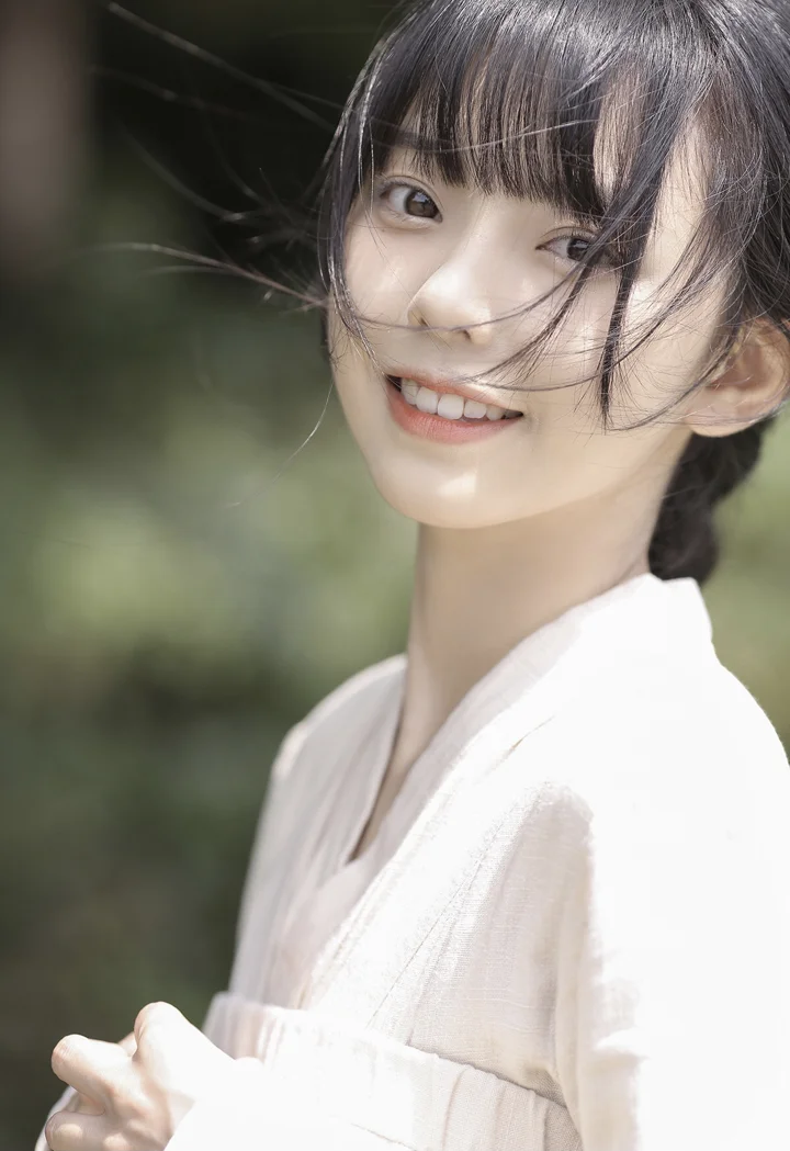 一位黑发女子穿着浅色上衣，在柔和的户外光线下微笑，背景是虚化的绿色植物。