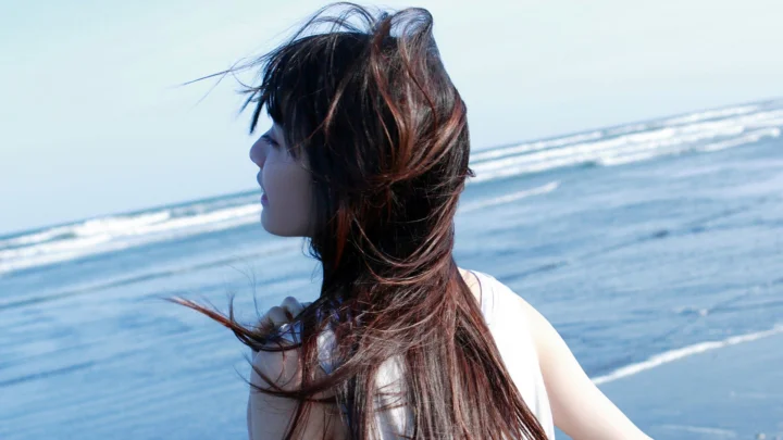 긴 머리의 여성이 바닷가에 서 있고 배경에는 파도와 푸른 바다가 펼쳐져 있습니다.