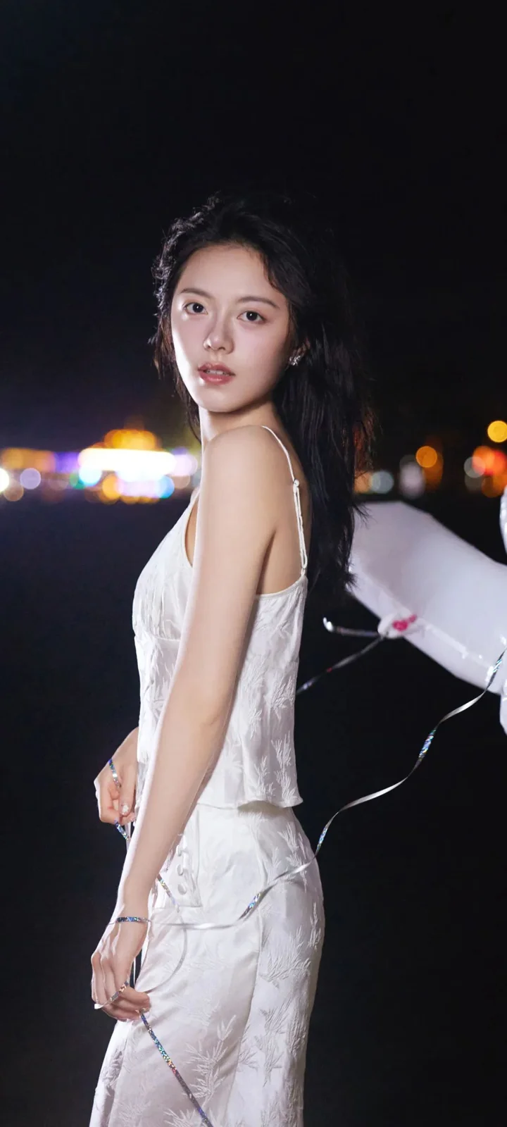 一名身穿白色连衣裙的女子在夜晚街头回望镜头，背景是柔和的城市灯光。
