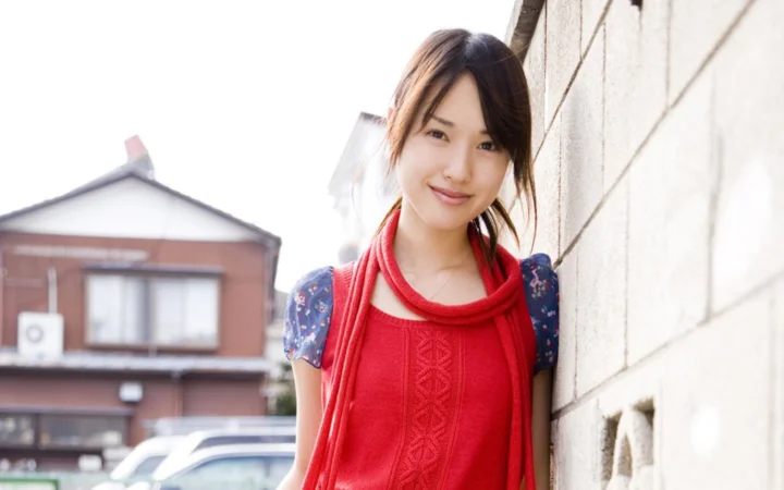 一名佩戴红色围巾的女子站在户外砖墙旁微笑面对镜头。