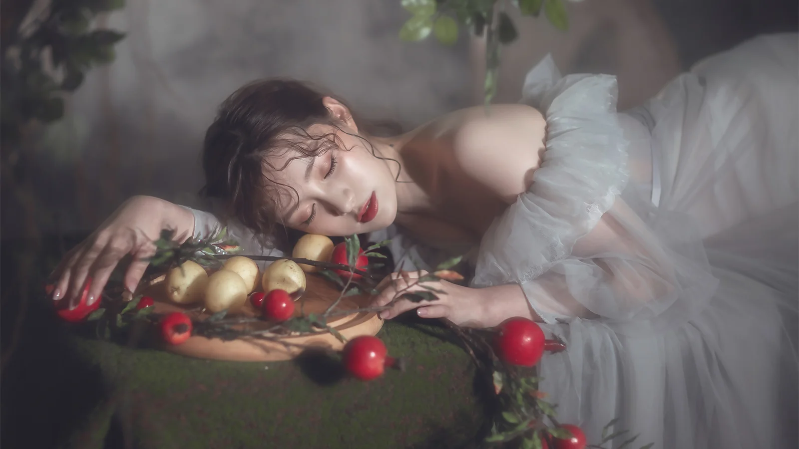 흰색 드레스를 입은 성인 여성이 초록색 천을 깐 테이블에 엎드려 있고 주변에 과일이 놓여 있습니다.