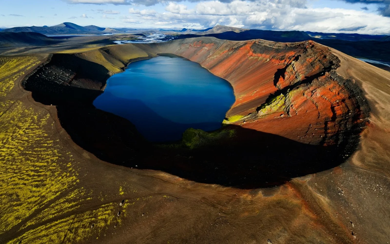 俯瞰一座深蓝色火山湖，四周环绕着黑色火山地貌和彩红色山坡，远处是群山与云层。