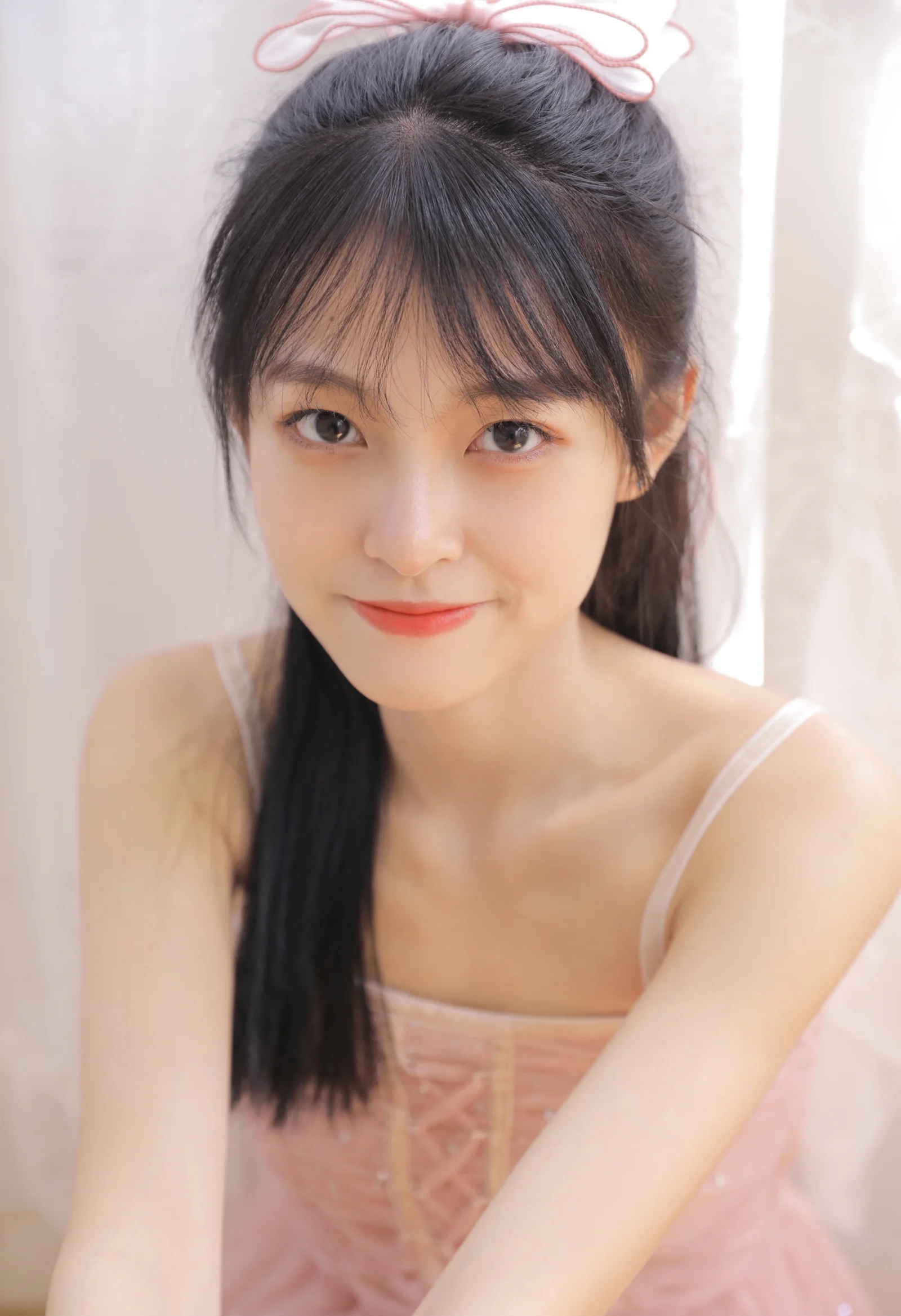 一位黑色長髮女性身穿粉色服飾，在柔和的室內光線下直視鏡頭微笑。