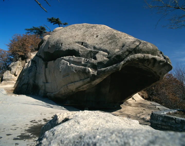 一块巨大的岩石悬于浅色岩地上方，周围点缀着树木，背景是晴朗的深蓝色天空。