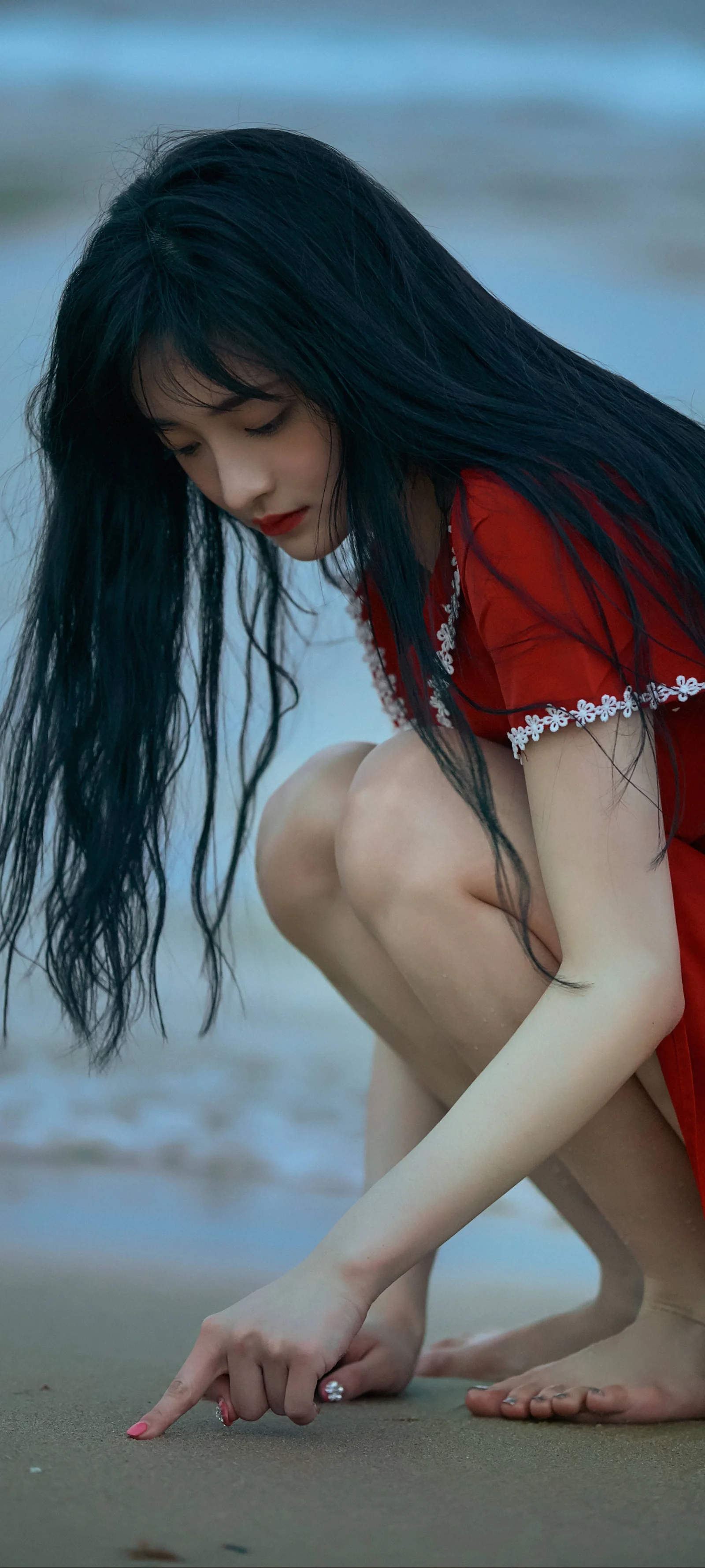長い黒髪の女性が赤いドレスを着て、海辺の濡れた砂の上にしゃがんでいます。