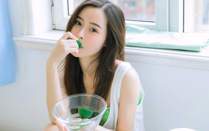 一名长发女子坐在窗边，手拿绿色水果并端着玻璃碗。