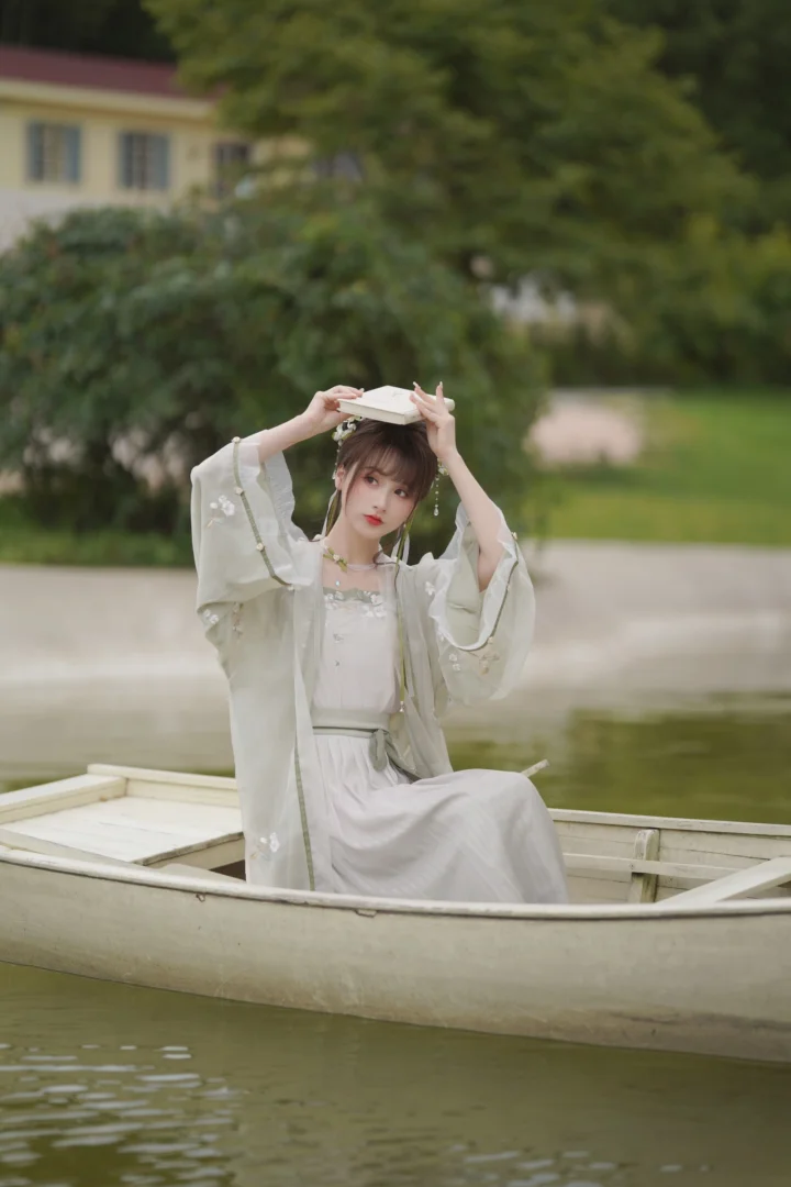 一位身穿浅色传统服饰的女子坐在湖面小船中，双手扶着头顶的白色物件。
