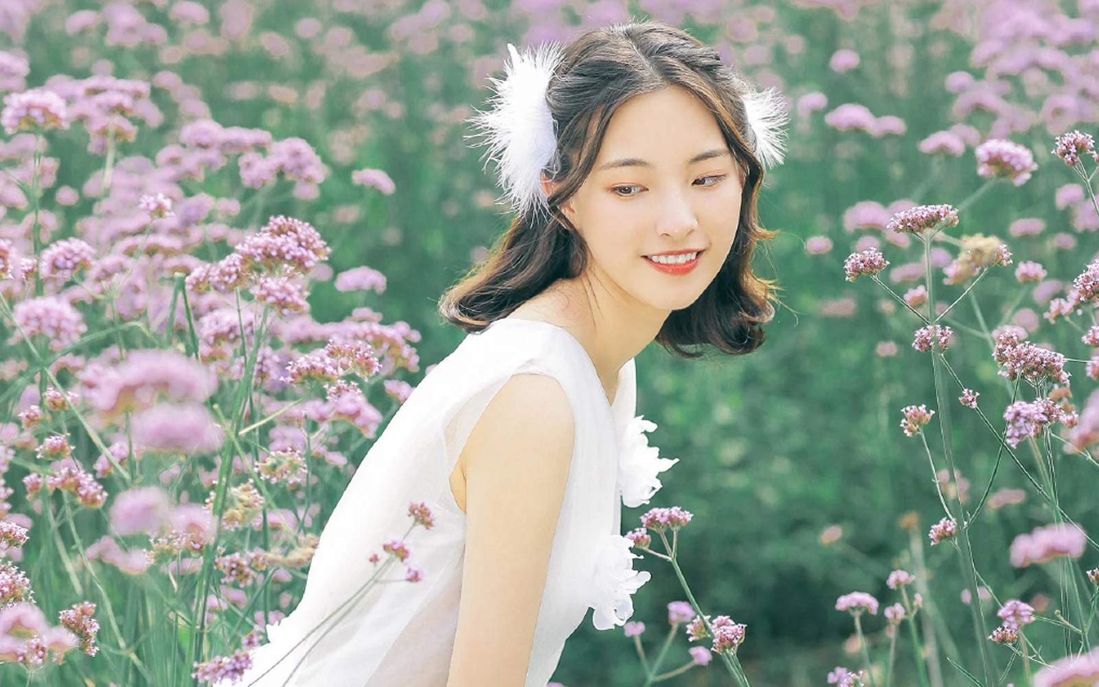 一名穿著白色無袖服裝的女子站在盛開的粉紫色花叢中。