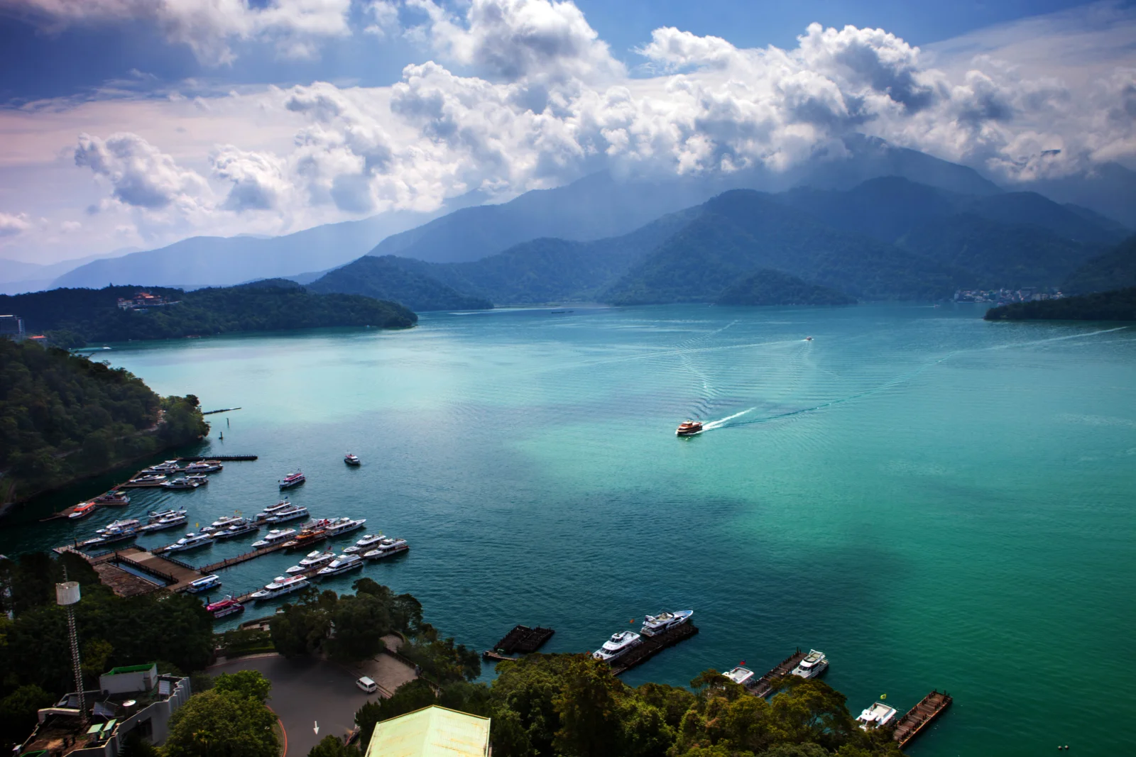 青緑色の湖が山々の間に広がり、空には幾重もの白い雲が浮かび、岸辺には小さな船が停泊しています。