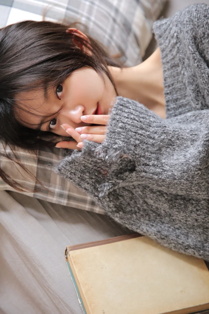 一名成年女性穿着灰色针织衫躺在格纹毯子上，旁边放着一本书。