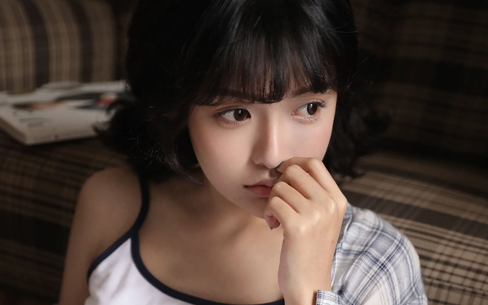 一名短发女生坐在室内沙发上，手靠近脸部并望向一侧。