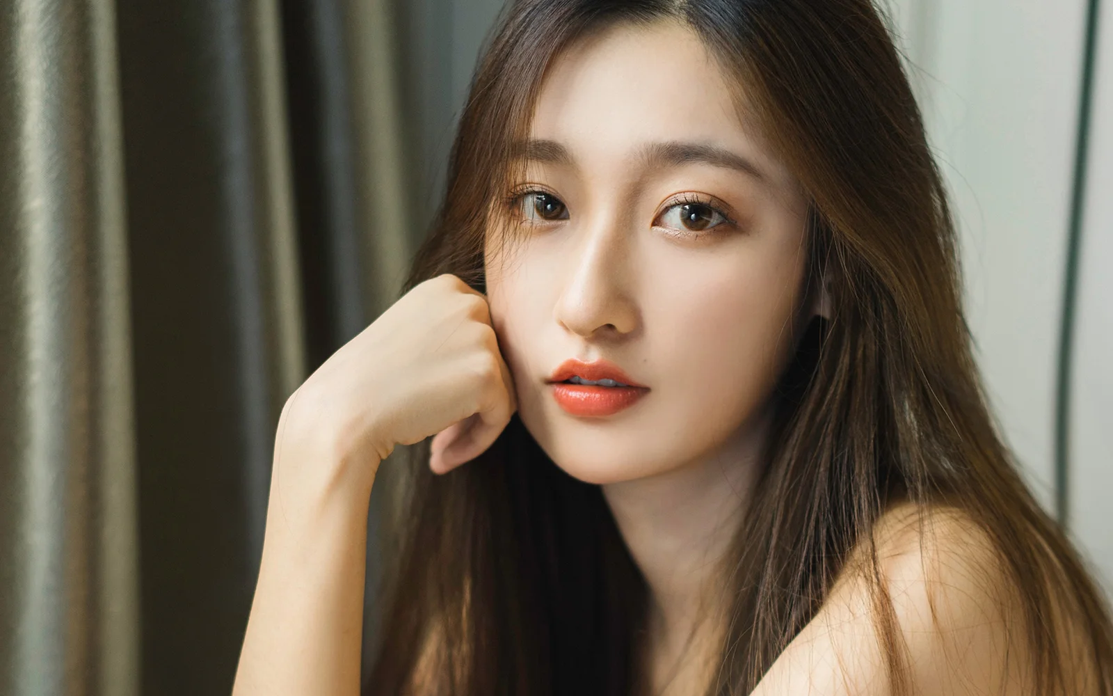 긴 머리의 여성이 손으로 얼굴을 받친 채 실내의 부드러운 조명 아래 카메라를 바라보고 있습니다.