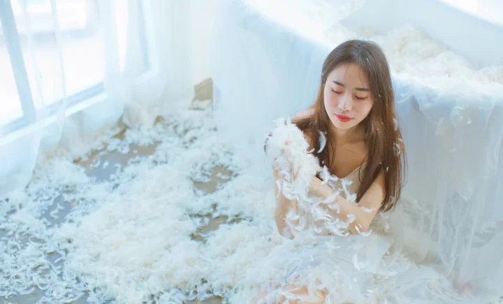 白い羽根の装飾が付いた服を着た成人女性が、明るい室内に座っています。
