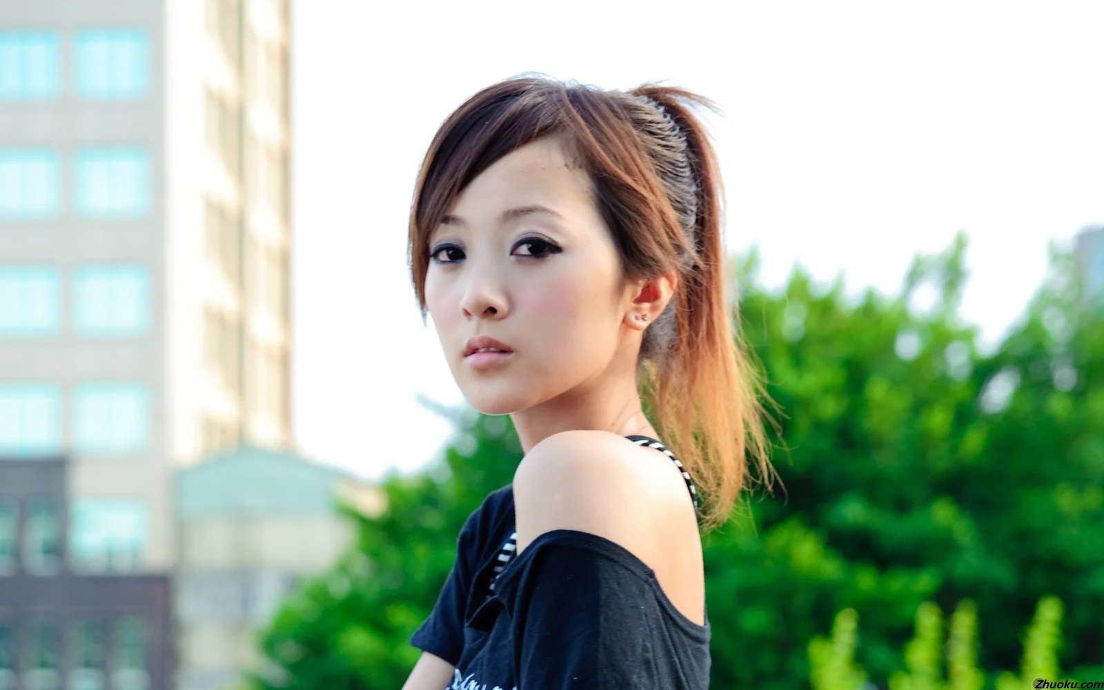 一名女性身穿黑色上衣在户外回望镜头，背景可见树木和建筑。