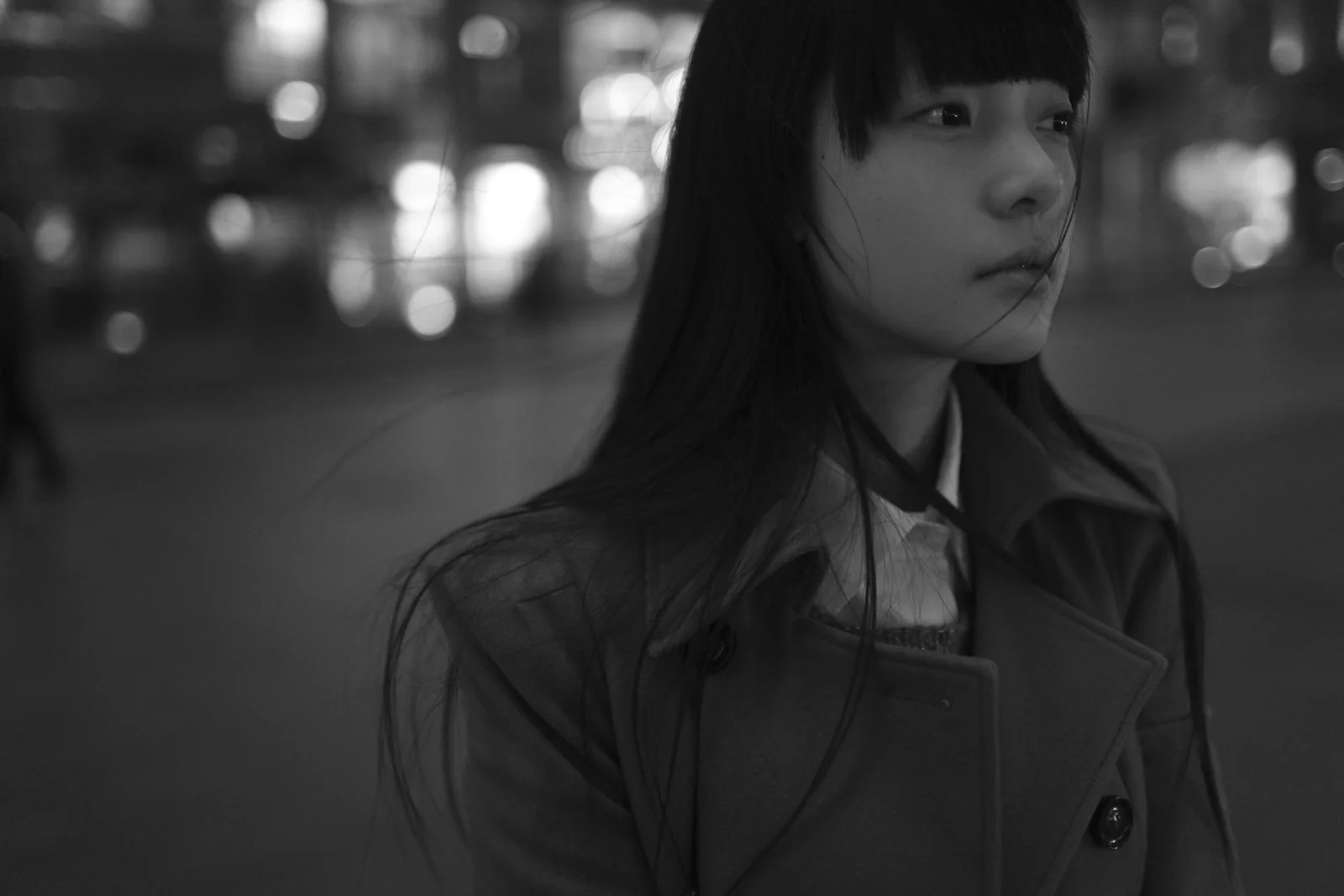 一名穿着深色外套的女性站在背景虚化的街道上，画面采用黑白色调。