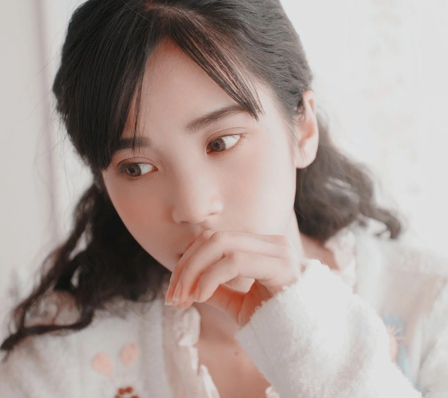 부드러운 빛 아래 흰색 니트 스웨터를 입은 성인 여성이 손을 입가에 댄 클로즈업 초상이다.