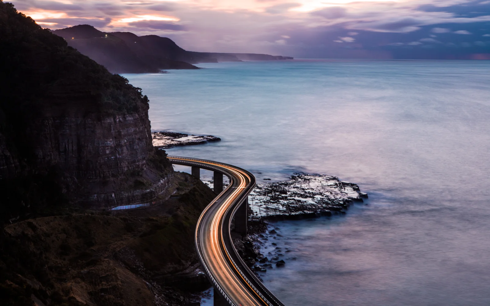 曲がりくねった海岸道路が険しい崖沿いに伸び、穏やかな海と遠くの山々が夕暮れの空の下に広がっています。