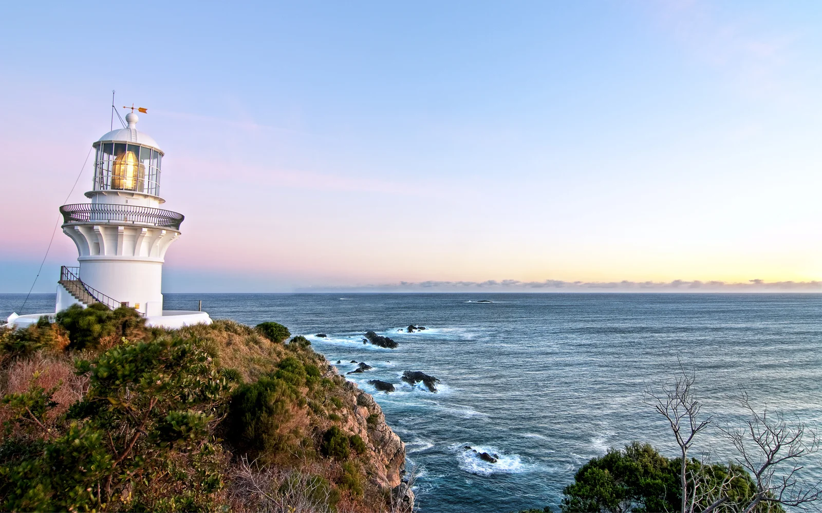 흰색 등대가 해안 절벽에 서서 옅은 파란색과 분홍색 하늘 아래 넓은 바다를 바라보고 있습니다.