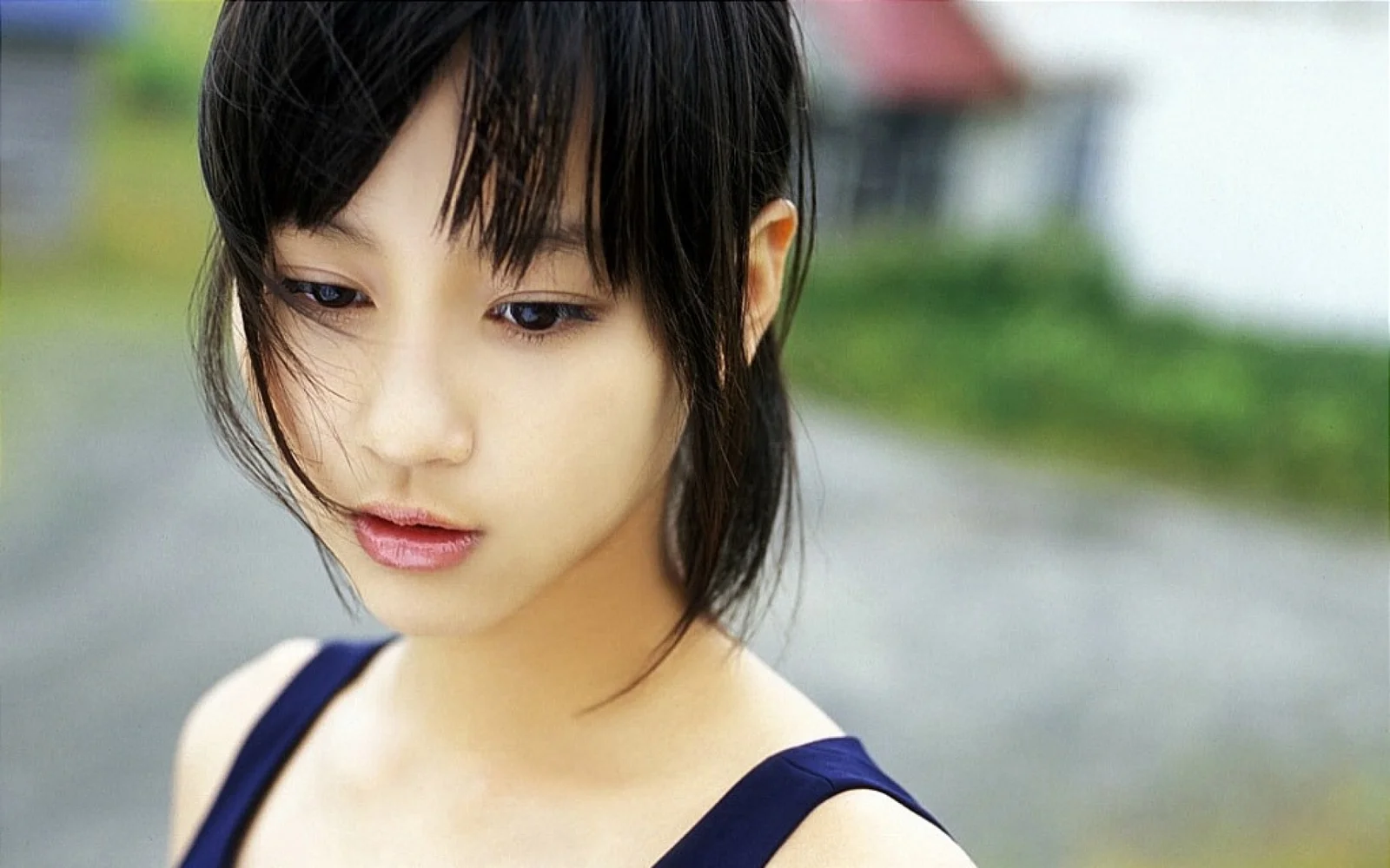 검은 머리의 여성이 흐릿한 야외 배경 앞에서 시선을 아래로 향하고 있습니다.
