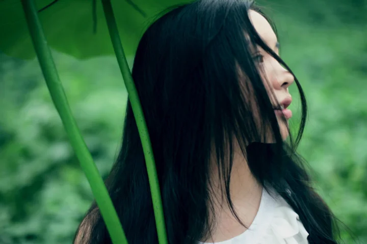 一名长黑发女子站在绿叶间，呈侧面姿态。